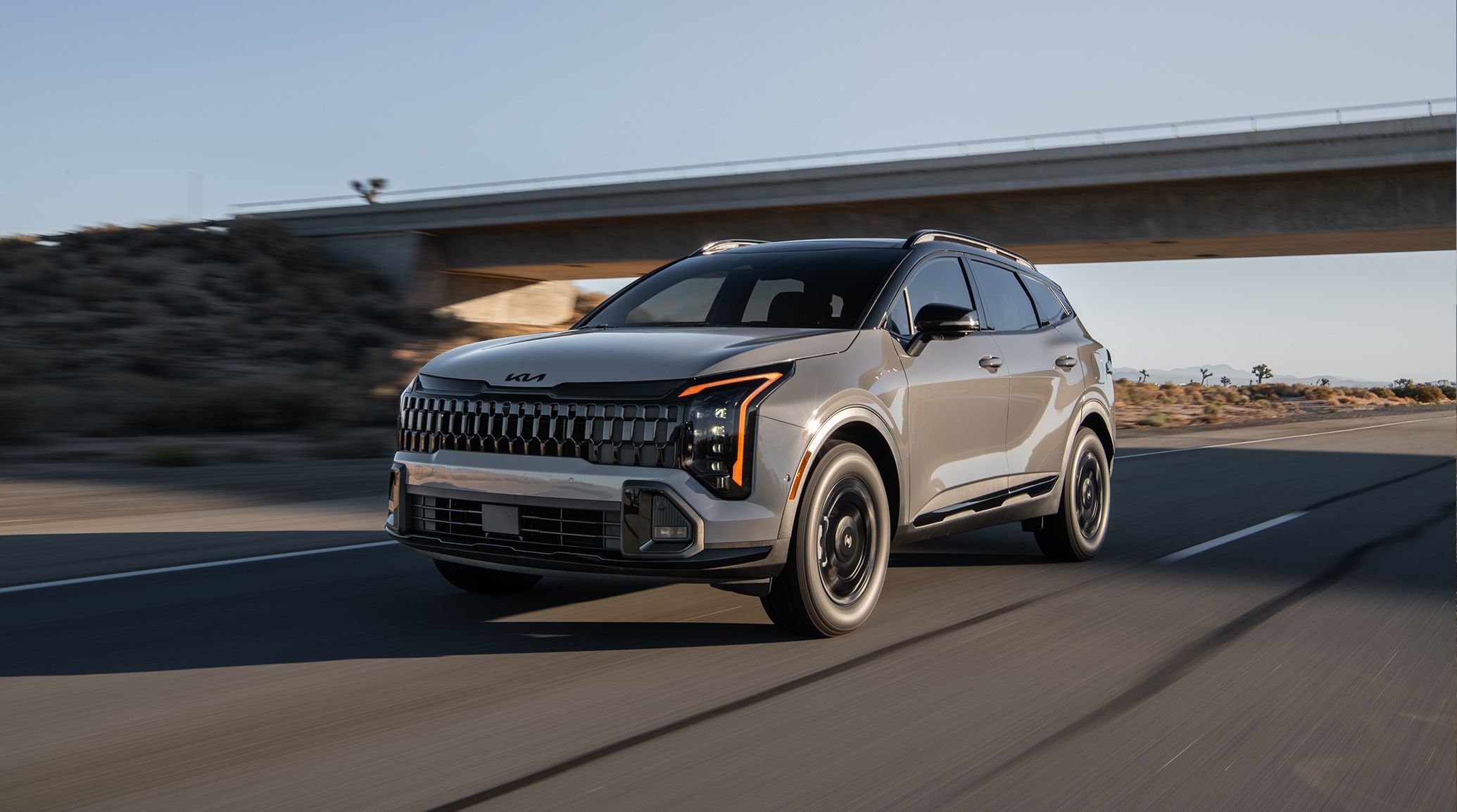 A light gray Kia Sportage driving past an overpass on a road in a desert