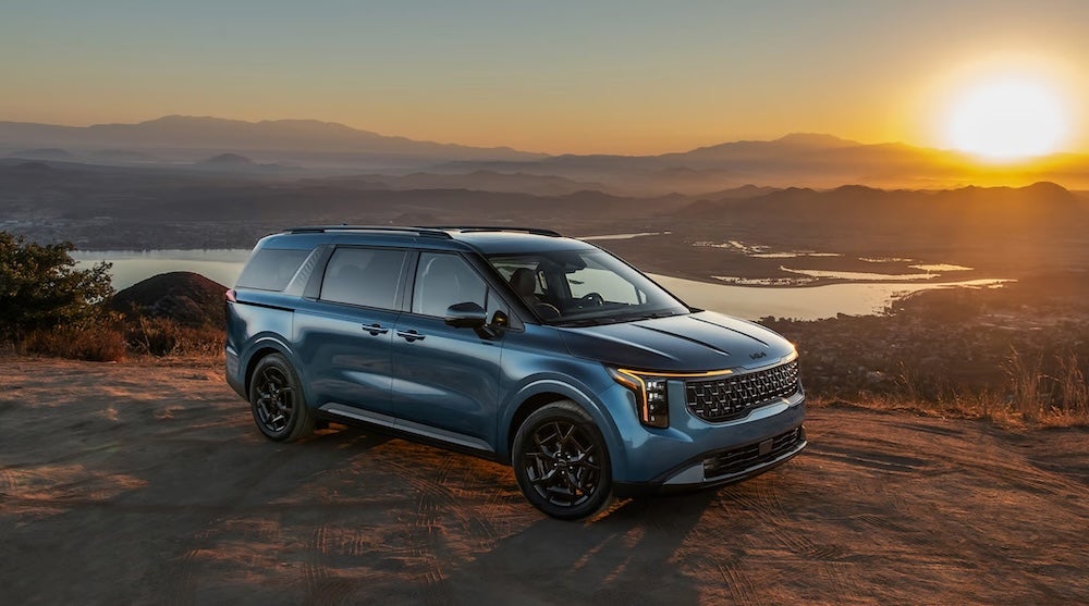 A blue 2027 Kia Carnival MPV parked on a ledge at sunset